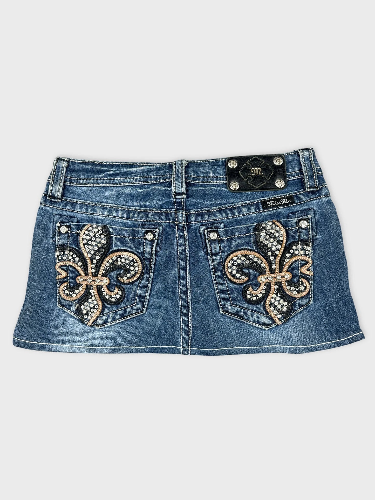 Back view of a blue denim skirt with embroidered fleur-de-lis patterns.